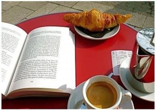 Sur une table en terrasse d'un café on aperçoit un livre ouvert, une tasse de café et un croissant.